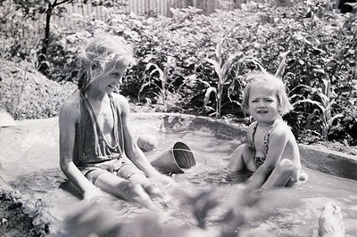 Two children play in a shallow outdoor pool surrounded by lush greenery, likely mid-20th century. The older child, seated, ho...