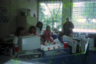 Vintage office scene with five seated individuals around a desk, likely late 1980s–early 1990s. Bulky CRT monitor, paper stac...
