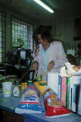 Indoor kitchen scene featuring a woman in a floral apron preparing food on a countertop. Visible items include a bag of potat...