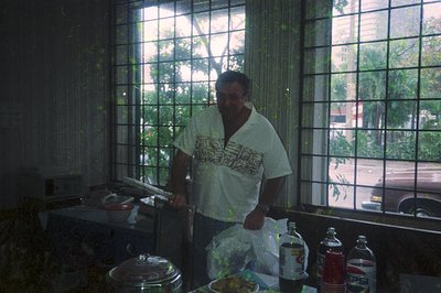 Indoor kitchen scene featuring a man in a white T-shirt with text, preparing food at a counter. Large grid-patterned window r...