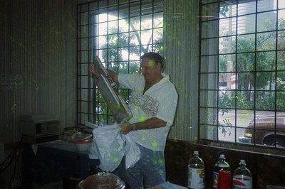 Man in a white apron with paint splatters operates a large spray gun indoors, likely painting or decorating. Surrounding item...