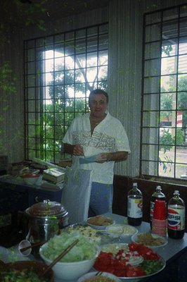 A man in a white apron stands in a bright kitchen, holding a menu or recipe card. Behind him, a large window frames lush gree...