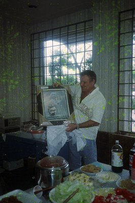 Man in 1970s-style apron holds framed photo in indoor kitchen, surrounded by food prep (bowls, pots, salad). Decor includes f...