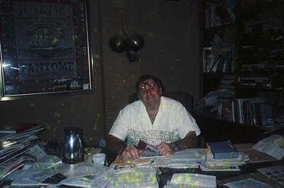 Vintage portrait of an elderly man seated at a cluttered desk, surrounded by stacks of papers, books, and framed posters. His...