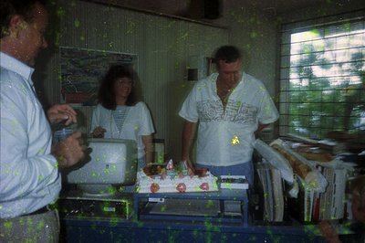 Indoor gathering with three adults preparing a decorated sheet cake on a metal table. Blurred vintage filter suggests low-res...