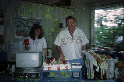Two adults pose indoors beside a birthday cake with "45" icing. The man wears a white shirt with a printed design and a lanya...