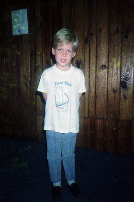 Young boy in a vintage "First Mate" T-shirt (1970s-era nautical theme) and faded jeans, standing indoors against a wooden wal...