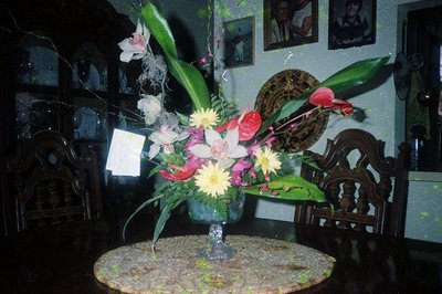 Vintage floral arrangement in a blue-green vase with orchids, daisies, and red accents on a round wooden table. Dark wooden c...