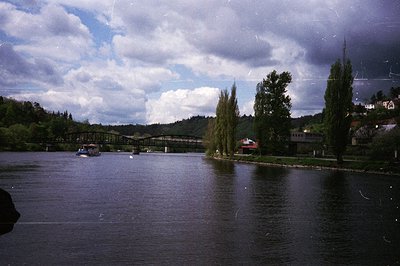 Vintage river scene with vintage bridge spanning calm waters, flanked by lush greenery and mature trees. Distant industrial-s...