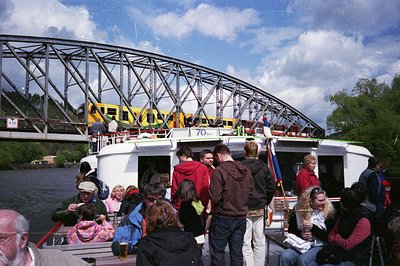 A vintage ferry boat transports passengers under a steel truss bridge marked "70," likely a 1970s-era structure. Crowds in ca...