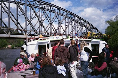A vintage riverboat labeled "70’s" transports a diverse crowd under a steel truss bridge, likely from the 1970s. The boat’s r...