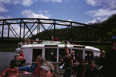 Vintage riverboat cruise with mid-century design, featuring a curved metal bridge in background. Crowded deck shows casual 19...