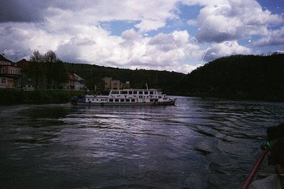 Passenger ferry navigating a wide river amid low-lying European town with brick buildings. Overcast skies and rippled water s...