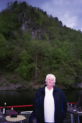 A mature individual stands on a wooden dock beside a serene lake, framed by dense forested hills. They wear a white shirt and...