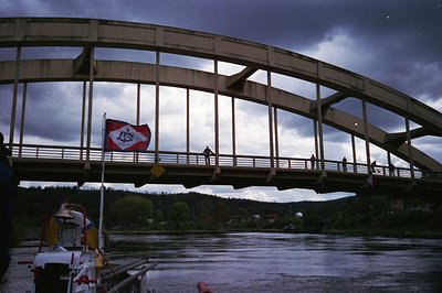 Concrete arch bridge spanning a river with industrial-style railings; flagpole displays a red/white emblem resembling a Sovie...