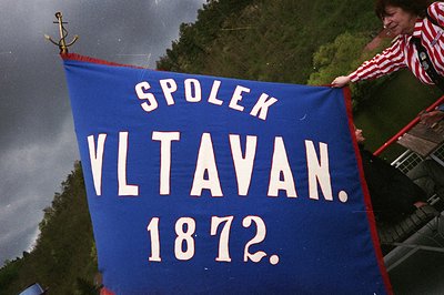 A person holds a vintage blue flag with white text reading "SPOLEK VLTAVAN. 1872," featuring a maritime anchor emblem at the ...