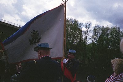 Military honor guard carrying a flag with a black eagle emblem on a red/white/blue background. Uniforms feature peaked caps a...