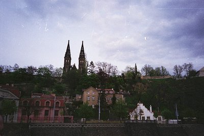Vintage black-and-white photo of a hillside town with Gothic Revival twin spires dominating skyline. Red-brick buildings with...