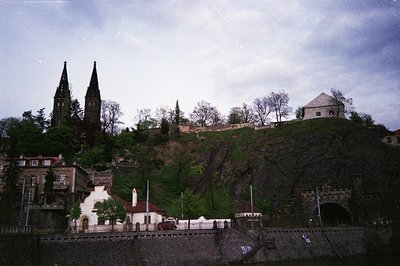 Historic hilltop fortress with twin spires and fortified walls, overlooking a riverbank town. Mid-20th century architecture b...