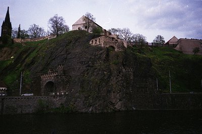 Historic stone fortress perched on rocky cliffside, featuring a prominent arched gateway and medieval architecture. Surroundi...