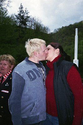 Two individuals share an affectionate moment outdoors, likely at an event or gathering. The man wears a gray hoodie with a vi...
