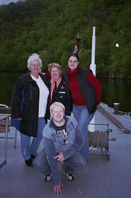 Four individuals pose on a wooden dock beside a serene lake, framed by lush greenery. The woman in the center wears a floral-...