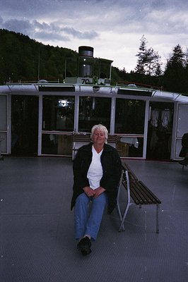 Retired individual seated on a vintage houseboat deck, numbered "70," with forest backdrop at dusk. Mid-20th century design f...