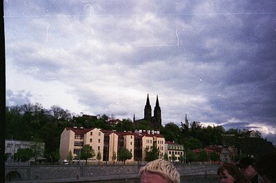 Vintage photo of a European riverside town with Gothic twin-spired church atop a hill. Mid-20th century architecture—brick bu...