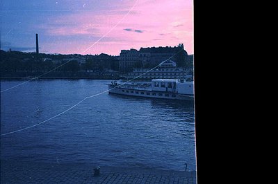Vintage urban riverscape at dusk, featuring a mid-20th-century ferry crossing with industrial chimneys and mid-rise residenti...