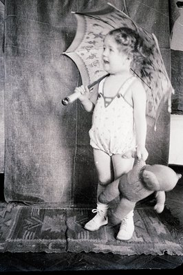 Vintage black-and-white photo of a young child playfully holding a decorative umbrella indoors, crouched near a tiled floor. ...