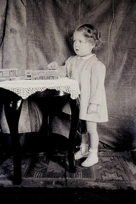 Vintage black-and-white portrait of a young girl (approx. 5-7 years) standing beside a wooden table in a studio setting. She ...