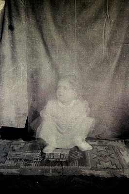 Vintage sepia-toned portrait of a child seated on ornate, patterned furniture, holding a small object. Studio backdrop featur...