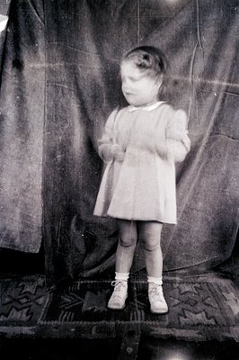 Vintage black-and-white portrait of a young child in a simple dress and white tights, standing on a patterned rug. Studio bac...