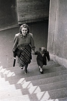 A woman in a plaid skirt and cardigan, holding a cane, descends concrete stairs with a child in a dark coat, mid-1950s. Archi...