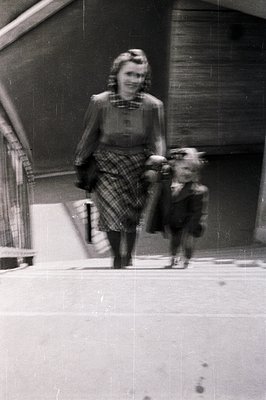 A woman in a 1950s-style dress and cardigan walks a small dog on a leash in an indoor setting with curved concrete walls. Her...