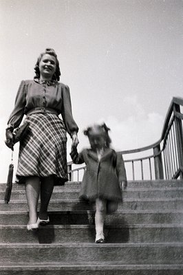 A mid-20th century black-and-white photo of a woman and child descending stone stairs. The woman wears a knee-length plaid sk...