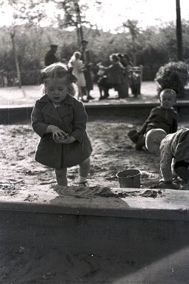 Black-and-white photo of children playing in a sandbox, likely mid-20th century. A toddler in a knee-length dress and hat kne...
