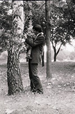 A man in 1950s-era suit and tie holds a young child outdoors, capturing a moment with a vintage camera. The scene features bi...
