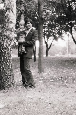Black-and-white portrait of a man in formal attire (vest, tie, suit) holding a young child outdoors, likely mid-20th century....