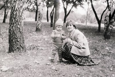 A mid-20th century black-and-white photo of a woman and toddler in a park setting. The woman, dressed in a cardigan, plaid sk...
