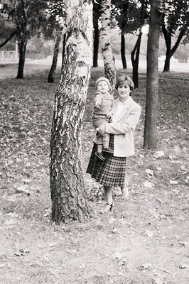 Black-and-white portrait of a woman cradling an infant in a park setting, likely mid-20th century. The woman wears a knee-len...
