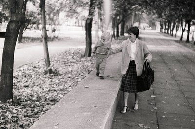 A mid-20th century black-and-white photo of a woman holding a toddler’s hands while walking on a tree-lined sidewalk. The wom...