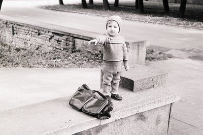 Toddler in vintage sweater and knit cap stands on concrete steps, pointing toward a large black leather satchel beside them. ...