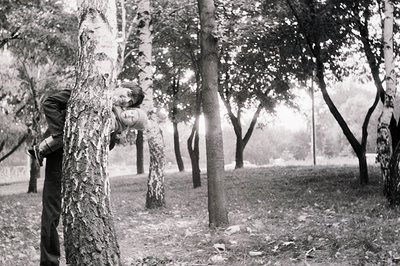 Black-and-white street scene: two individuals embrace under a tree in a park, likely mid-20th century. Urban greenery with ma...