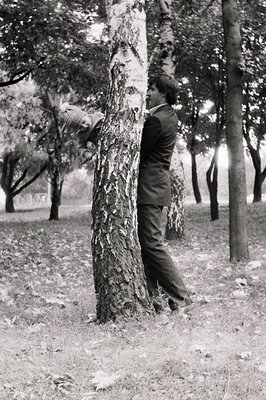Black-and-white portrait of a man in 1930s-40s formalwear leaning against a tree trunk in a park setting. His posture suggest...