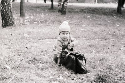 Mid-20th century black-and-white photo of a child sitting on dry grass in a park, holding a vintage leather suitcase. Cozy kn...