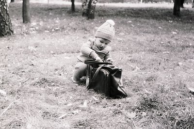Vintage black-and-white photo of a toddler in winter attire—knit hat and plaid jacket—squatting on dry grass to examine a bro...