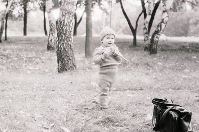 Black-and-white photo of a toddler in outdoor park setting, mid-stride with a stick. Wearing a knitted hat and long-sleeve sw...