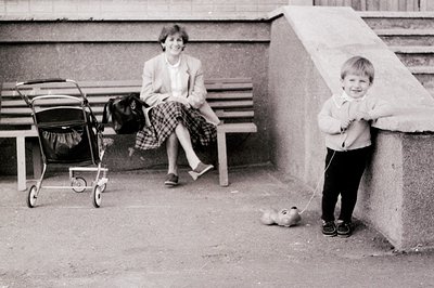 A 1970s-era black-and-white photo captures a candid urban moment: a woman in a blazer and plaid skirt sits on concrete steps ...