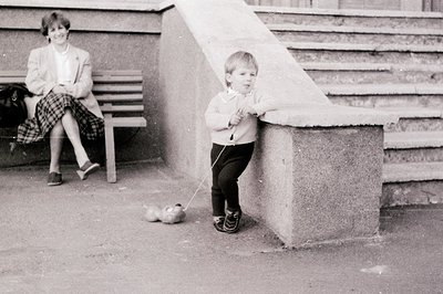 A young child in 1970s-era clothing holds a leash while walking a small dog beside a concrete staircase. An adult sits on a b...